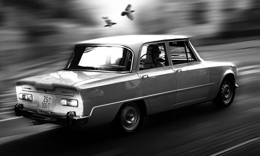 A black and white photograph of a vintage car speeding on a highway with bird flying next to it with a blurred background.