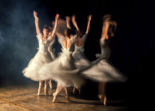 A photograph of five ballerinas in white tutus performing on stage, captured with a slow shutter speed to create motion blur.